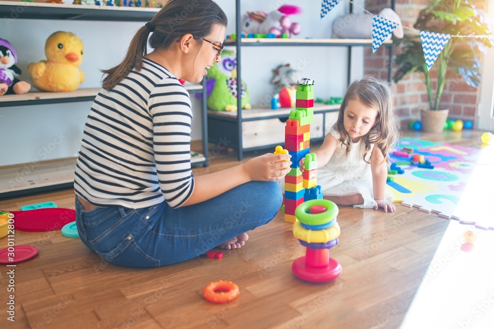 Fototapeta premium Beautiful teacher woman and toddler playing with building blocks around lots of toys at kindergarten