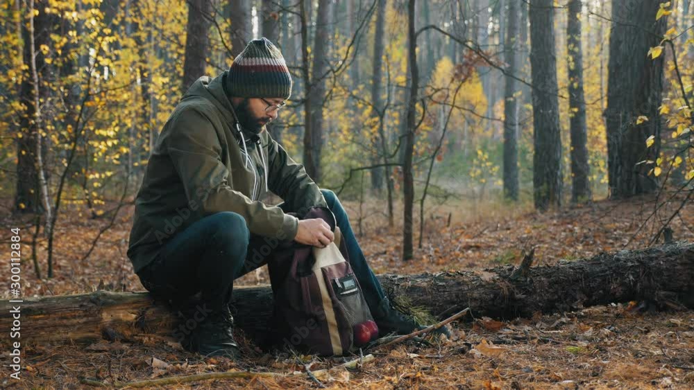 A bearded guy dressed in warm clothes sits on a log in the autumn forest, puts a thermos in his backpack and goes deep into the forest A bearded guy dressed in warm clothes sits on a log in the autumn forest, puts a thermos in his backpack and goes deep into the forest
