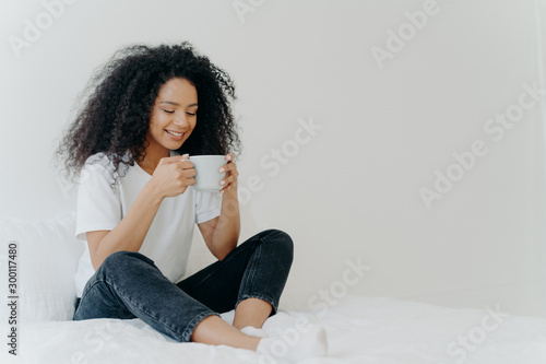 Photography African American woman sips hot drink, poses in bed, enjoys good morning, dressed casually, spends weekend at home, blank space aside for your advertising