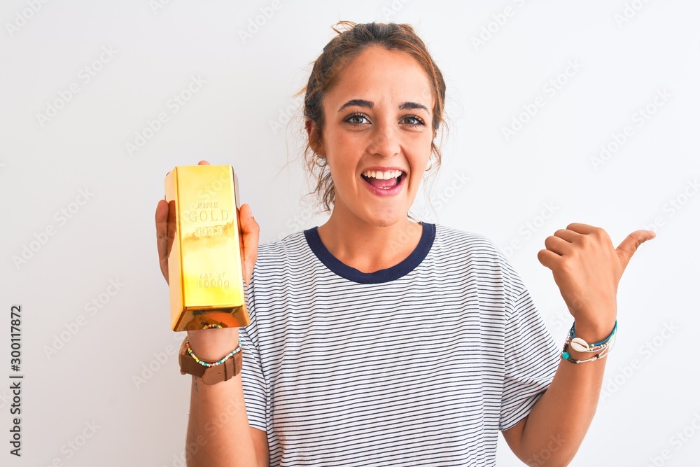 Young beautiful redhead woman holding gold ingot over isolated background pointing and showing with thumb up to the side with happy face smiling Young beautiful redhead woman holding gold ingot over isolated background pointing and showing with thumb up to the side with happy face smiling