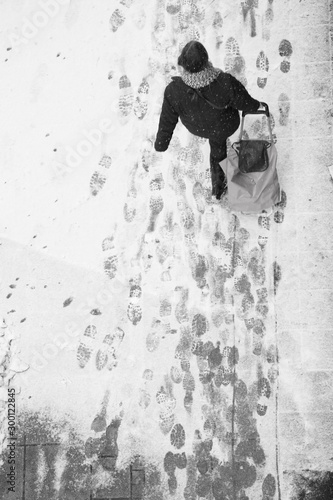 Fototapeta Naklejka Na Ścianę i Meble -  Blurry woman walking while pulling a shopping trolley bag during snowfall, high angle view
