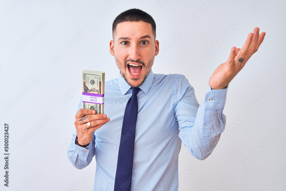 Young handsome business man holding a bunch of dollars bank notes over isolated background very happy and excited, winner expression celebrating victory screaming with big smile and raised hands Young handsome business man holding a bunch of dollars bank notes over isolated background very happy and excited, winner expression celebrating victory screaming with big smile and raised hands