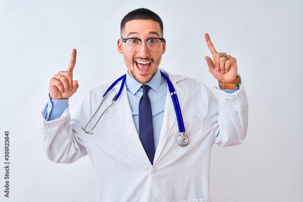 Young doctor man wearing stethoscope over isolated background smiling amazed and surprised and pointing up with fingers and raised arms. Young doctor man wearing stethoscope over isolated background smiling amazed and surprised and pointing up with fingers and raised arms.