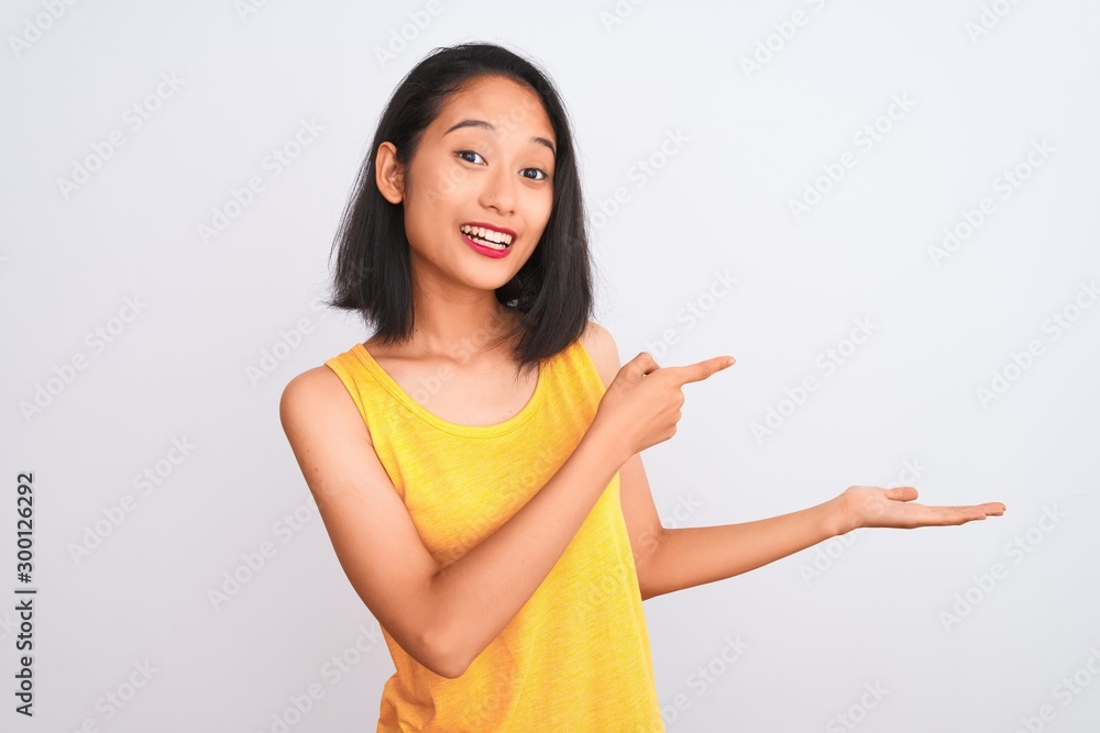 Young chinese woman wearing yellow casual t-shirt standing over isolated white background amazed and smiling to the camera while presenting with hand and pointing with finger. Young chinese woman wearing yellow casual t-shirt standing over isolated white background amazed and smiling to the camera while presenting with hand and pointing with finger.