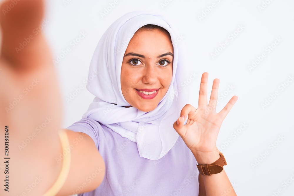 Beautiful arabian girl wearing hijab make selfie by camera over isolated white background doing ok sign with fingers, excellent symbol Beautiful arabian girl wearing hijab make selfie by camera over isolated white background doing ok sign with fingers, excellent symbol
