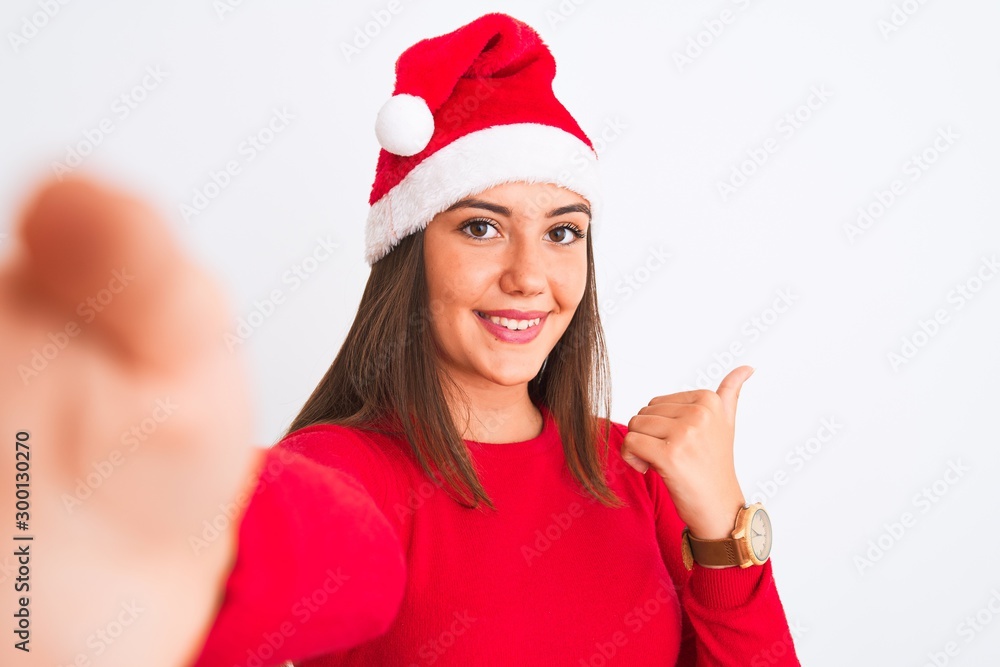 Girl wearing Christmas Santa hat make selfie by camera over isolated white background pointing and showing with thumb up to the side with happy face smiling Girl wearing Christmas Santa hat make selfie by camera over isolated white background pointing and showing with thumb up to the side with happy face smiling