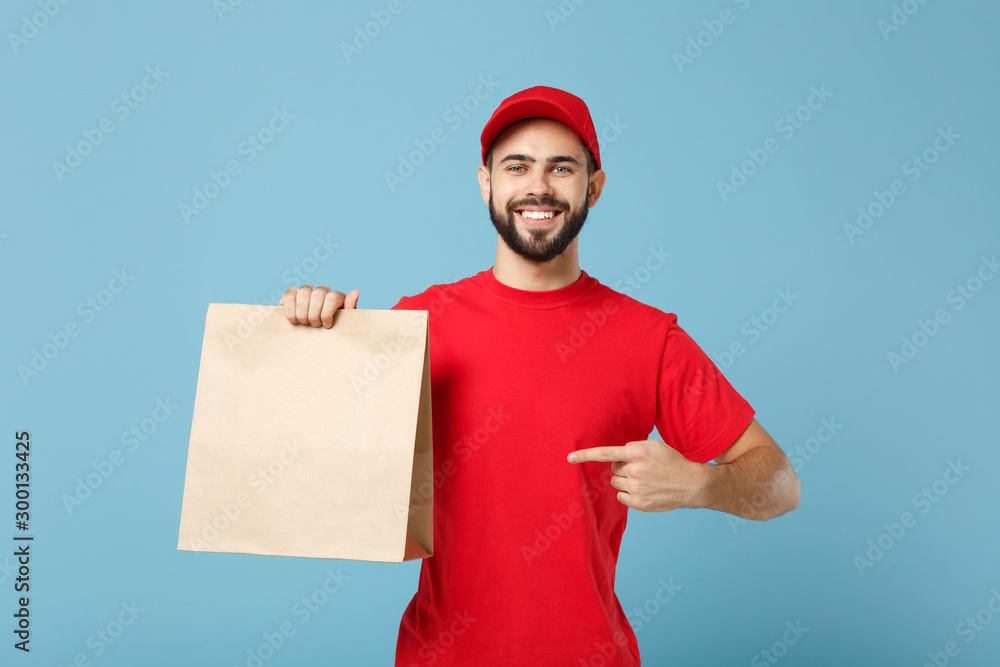 Delivery man in red uniform hold craft paper packet with food isolated on blue background, studio portrait. Male employee in cap t-shirt print working as courier. Service concept. Mock up copy space. Delivery man in red uniform hold craft paper packet with food isolated on blue background, studio portrait. Male employee in cap t-shirt print working as courier. Service concept. Mock up copy space.