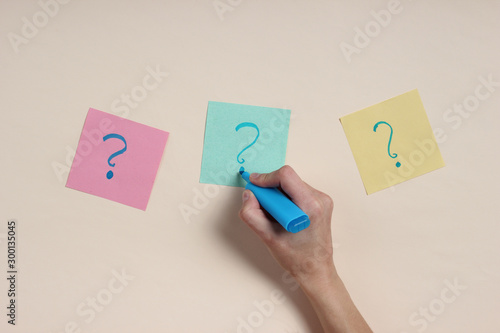 Female hand holds a blue felt-tip pen and draws question marks on memo papers against beige background. Top view