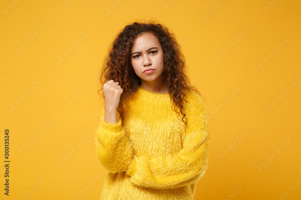Angry strict young african american girl in fur sweater posing isolated on yellow orange background, studio portrait. People sincere emotions lifestyle concept. Mock up copy space. Clenching fists. Angry strict young african american girl in fur sweater posing isolated on yellow orange background, studio portrait. People sincere emotions lifestyle concept. Mock up copy space. Clenching fists.