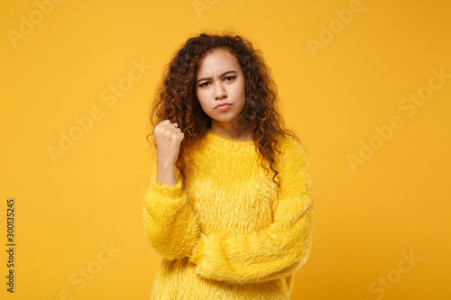 Angry strict young african american girl in fur sweater posing isolated on yellow orange background, studio portrait. People sincere emotions lifestyle concept. Mock up copy space. Clenching fists. © ViDi Studio