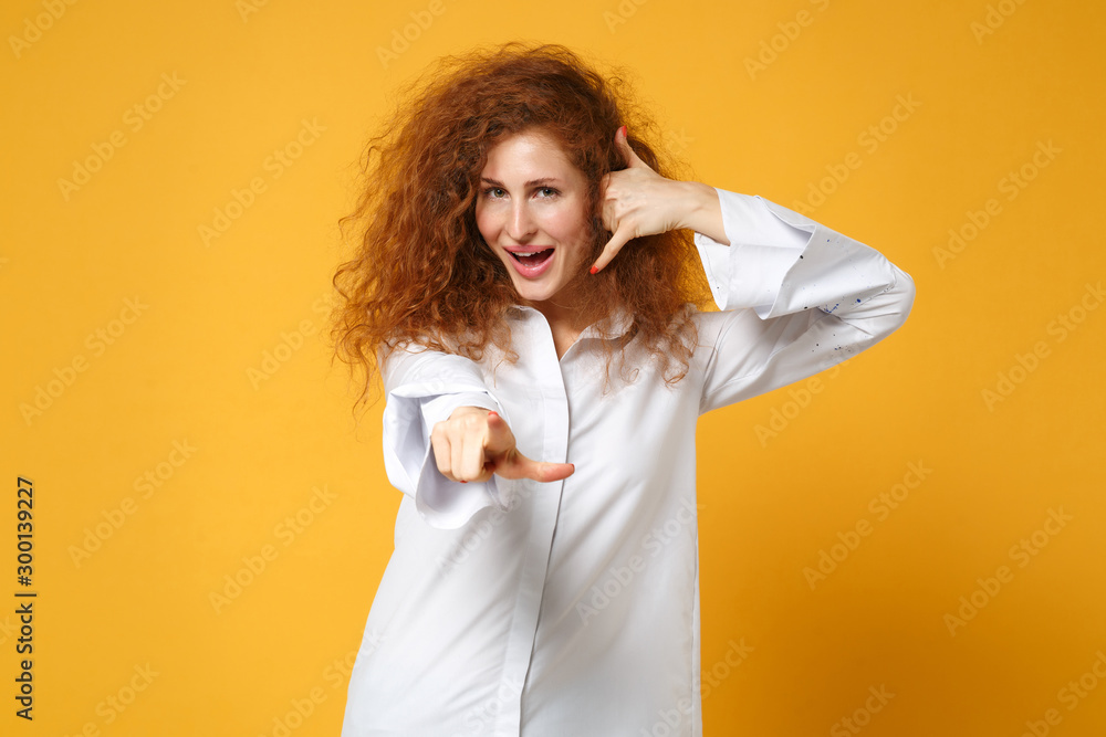 Young redhead woman girl in shirt posing isolated on yellow orange background. People lifestyle concept. Mock up copy space. Doing phone gesture like says call me back pointing index finger on camera. Young redhead woman girl in shirt posing isolated on yellow orange background. People lifestyle concept. Mock up copy space. Doing phone gesture like says call me back pointing index finger on camera.