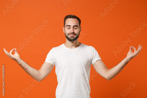 Young man in casual white t-shirt posing isolated on orange background in studio. People lifestyle concept. Mock up copy space. Hold hands in yoga gesture, relaxing meditating, keeping eyes closed.