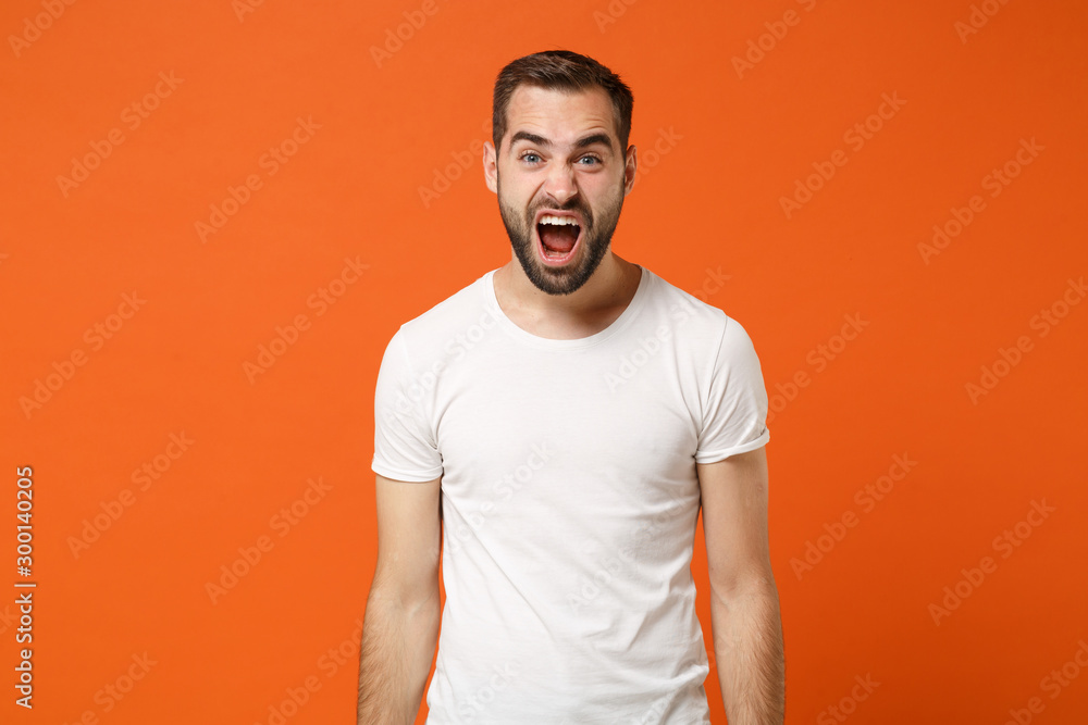 Angry crazy young man in casual white t-shirt posing isolated on bright orange wall background, studio portrait. People sincere emotions lifestyle concept. Mock up copy space. Screaming, swearing. Angry crazy young man in casual white t-shirt posing isolated on bright orange wall background, studio portrait. People sincere emotions lifestyle concept. Mock up copy space. Screaming, swearing.