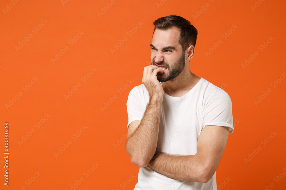 Nervous young man in casual white t-shirt posing isolated on bright orange wall background studio portrait. People sincere emotions lifestyle concept. Mock up copy space. Gnawing nails, looking aside. Nervous young man in casual white t-shirt posing isolated on bright orange wall background studio portrait. People sincere emotions lifestyle concept. Mock up copy space. Gnawing nails, looking aside.