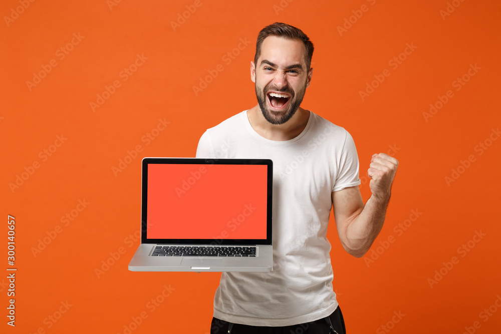 Excited young man in casual white t-shirt posing isolated on orange background. People lifestyle concept. Mock up copy space. Holding laptop pc computer with blank empty screen, doing winner gesture. Excited young man in casual white t-shirt posing isolated on orange background. People lifestyle concept. Mock up copy space. Holding laptop pc computer with blank empty screen, doing winner gesture.