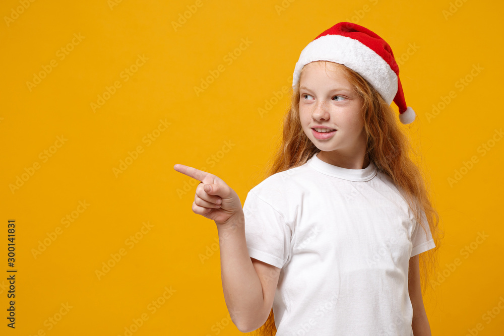 Little ginger kid Santa girl 12-13 years old in white t-shirt Christmas hat isolated on yellow background. New Year 2020 celebration holiday concept. Mock up copy space. Pointing index finger aside. Little ginger kid Santa girl 12-13 years old in white t-shirt Christmas hat isolated on yellow background. New Year 2020 celebration holiday concept. Mock up copy space. Pointing index finger aside.