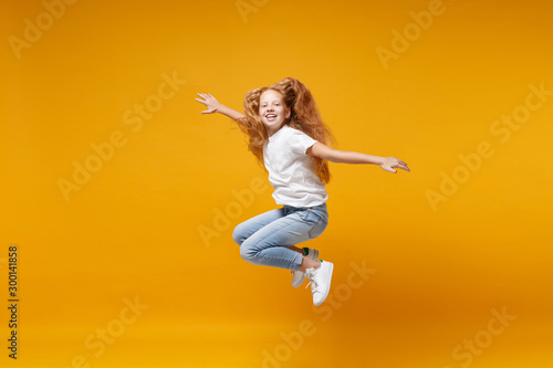 Pretty little ginger kid girl 12-13 years old in white t-shirt isolated on yellow background. Childhood lifestyle concept. Mock up copy space. Having fun, fooling around, jumping, spreading hands.