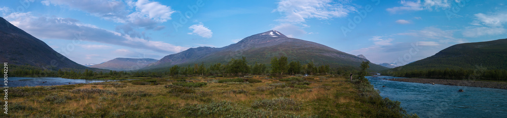 Wide panoramatic Lapland nature landscape with angler fisherman and small tent at blue Tjaktjajakka river, Kaitumjaure, birch tree forest and mountain Sanjartjakka. Summer at North sweden wilderness.