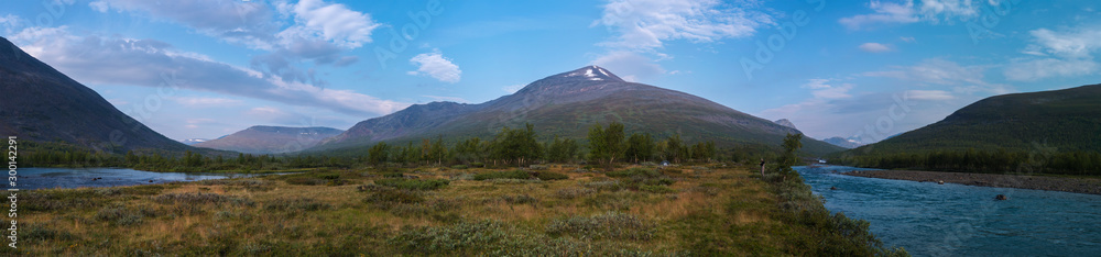 Wide panoramatic Lapland nature landscape with angler fisherman and small tent at blue Tjaktjajakka river, Kaitumjaure, birch tree forest and mountain Sanjartjakka. Summer at North sweden wilderness. Wide panoramatic Lapland nature landscape with angler fisherman and small tent at blue Tjaktjajakka river, Kaitumjaure, birch tree forest and mountain Sanjartjakka. Summer at North sweden wilderness.