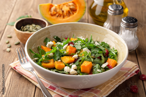 Autumn salad with baked pumpkin, arugula, seeds, dried cranberries and feta cheese in bowl on rustic wooden background. Selective focus.