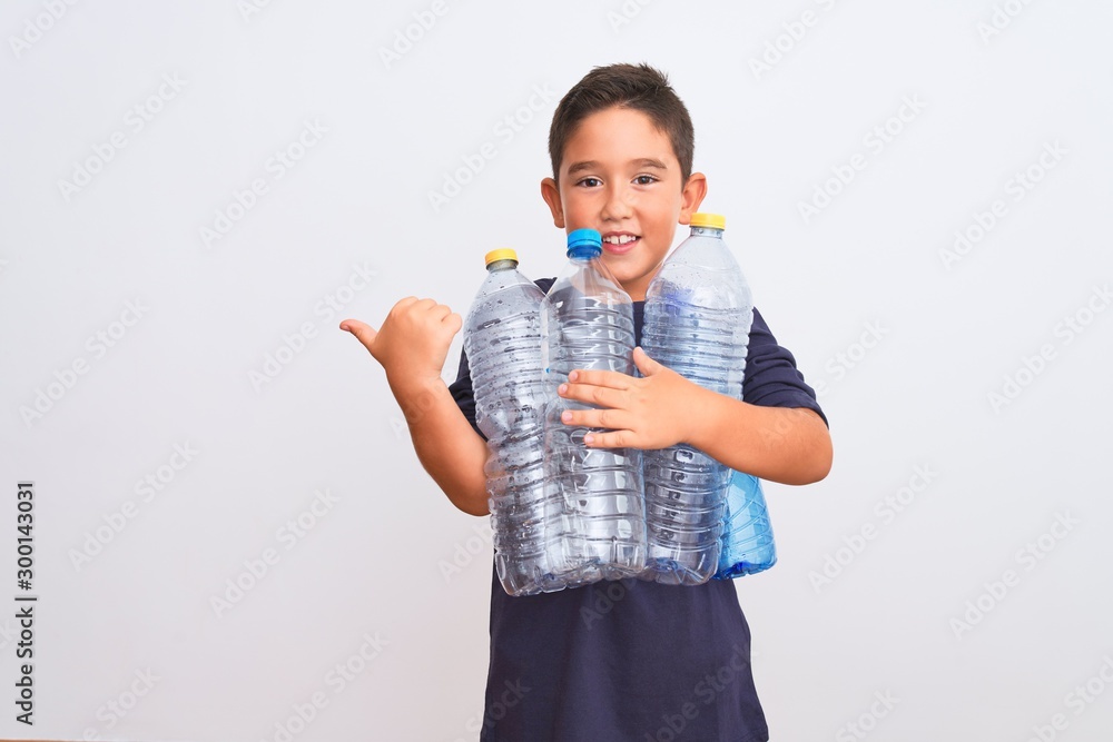 Beautiful kid boy recycling plastic bottles standing over isolated white background pointing and showing with thumb up to the side with happy face smiling Beautiful kid boy recycling plastic bottles standing over isolated white background pointing and showing with thumb up to the side with happy face smiling