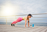 Young fit woman in sportswear and cap doing push ups exercises on the beach in the morning
