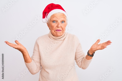 Senior grey-haired woman wearing Crhistmas Santa hat over isolated white background clueless and confused expression with arms and hands raised. Doubt concept.