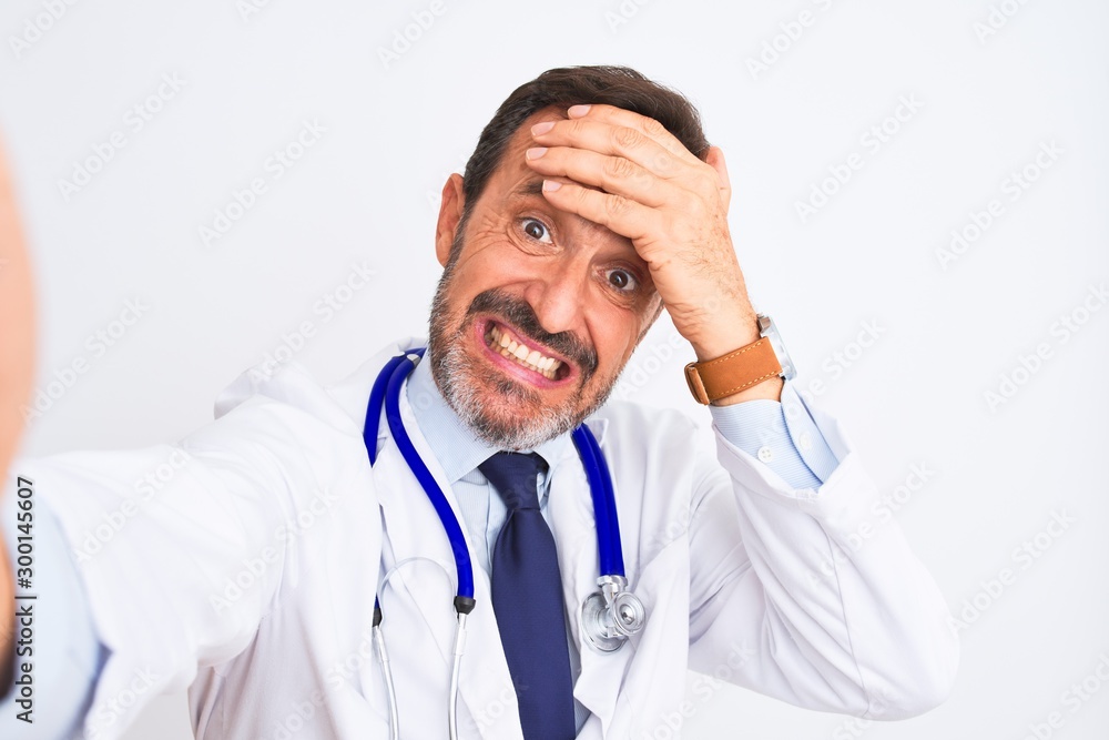 Middle age doctor man wearing stethoscope make selfie over isolated white background stressed with hand on head, shocked with shame and surprise face, angry and frustrated. Fear and upset for mistake. Middle age doctor man wearing stethoscope make selfie over isolated white background stressed with hand on head, shocked with shame and surprise face, angry and frustrated. Fear and upset for mistake.