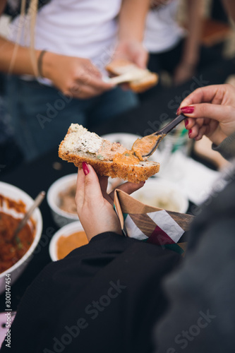 Woman hands holds chickpea hummus on spoon. The woman puts the hummus on a slice of bread. Outside party. Another person and bowls of hummus in the background. Close up
