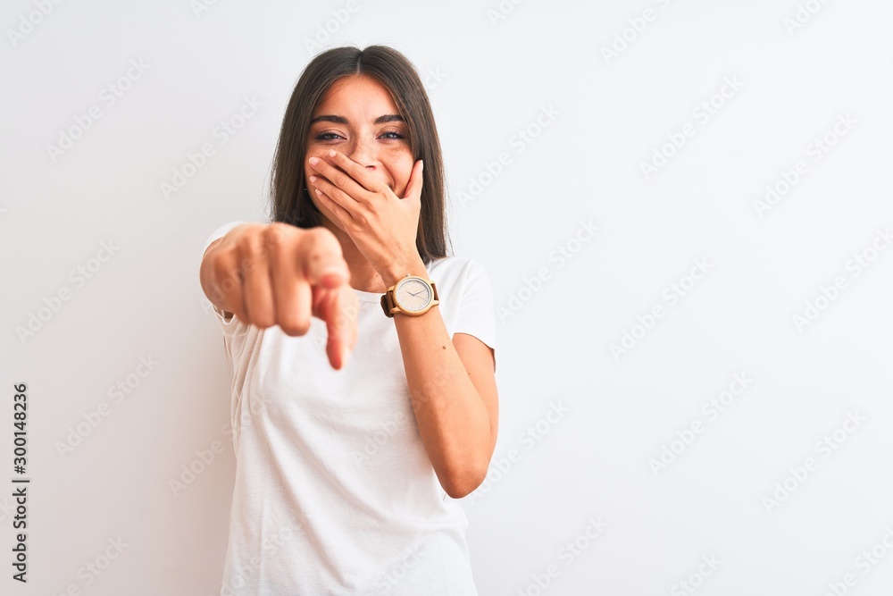 Young beautiful woman wearing casual t-shirt standing over isolated white background laughing at you, pointing finger to the camera with hand over mouth, shame expression Young beautiful woman wearing casual t-shirt standing over isolated white background laughing at you, pointing finger to the camera with hand over mouth, shame expression