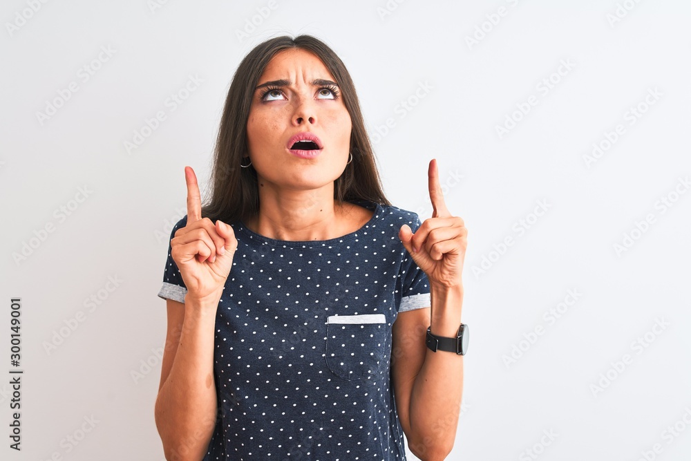 Young beautiful woman wearing blue casual t-shirt standing over isolated white background amazed and surprised looking up and pointing with fingers and raised arms. Young beautiful woman wearing blue casual t-shirt standing over isolated white background amazed and surprised looking up and pointing with fingers and raised arms.