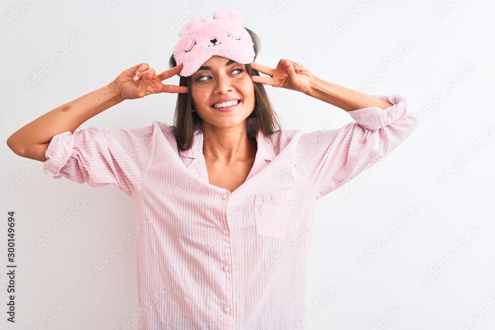 Young beautiful woman wearing sleep mask and pajama over isolated white background Doing peace symbol with fingers over face, smiling cheerful showing victory Young beautiful woman wearing sleep mask and pajama over isolated white background Doing peace symbol with fingers over face, smiling cheerful showing victory