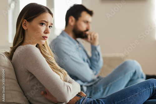 Worried young woman sitting on sofa at home and ignoring her boyfriend who is sitting next to her photo
