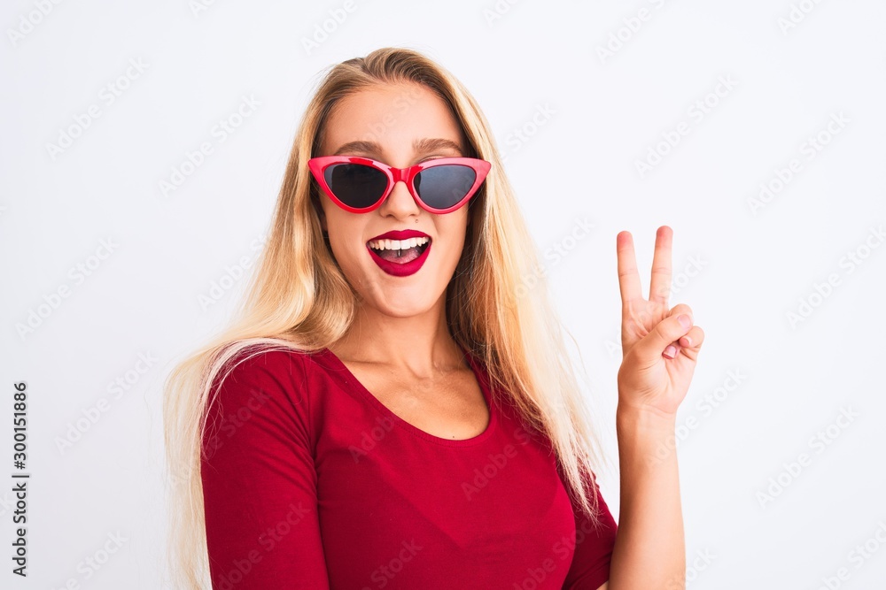 Naklejka premium Young beautiful woman wearing red t-shirt and sunglasses over isolated white background smiling with happy face winking at the camera doing victory sign. Number two.