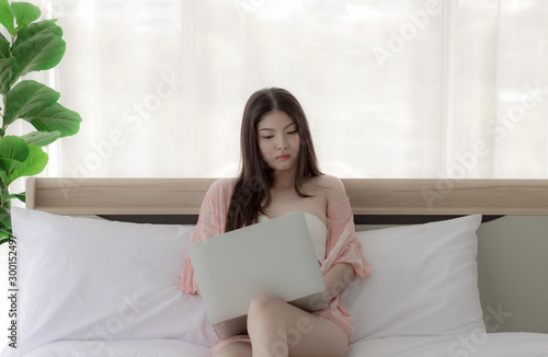 Asian woman wearing pink dress sitting on white couch. In a clean white bedroom woman sitting and working using notebook computer to communicate about business. The current purchase is convenient.