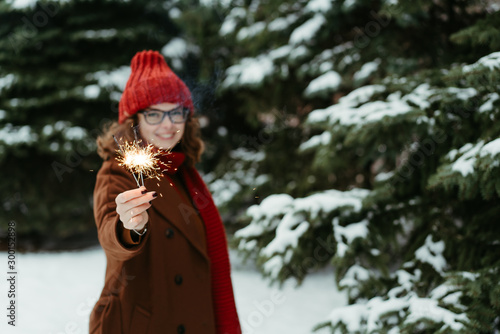 Young beautiful woman in knitted red hat and scarf standing in the park with bengal light, sparkler. Concept celebration, christmas