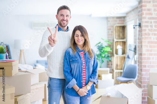 Young beautiful couple standing at new home around cardboard boxes showing and pointing up with fingers number two while smiling confident and happy.