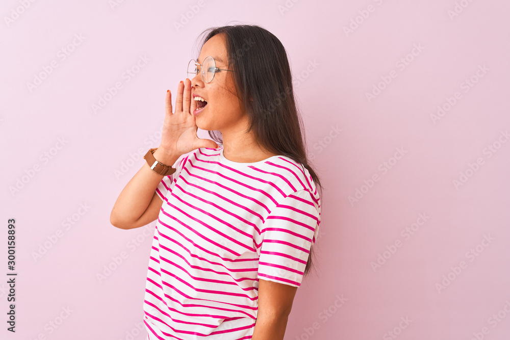 Young chinese woman wearing striped t-shirt and glasses over isolated pink background shouting and screaming loud to side with hand on mouth. Communication concept. Young chinese woman wearing striped t-shirt and glasses over isolated pink background shouting and screaming loud to side with hand on mouth. Communication concept.