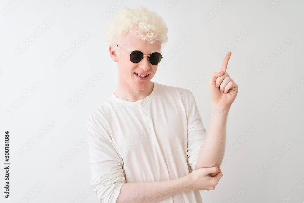 Young albino blond man wearing t-shirt and sunglasses over isolated white background with a big smile on face, pointing with hand and finger to the side looking at the camera. Young albino blond man wearing t-shirt and sunglasses over isolated white background with a big smile on face, pointing with hand and finger to the side looking at the camera.