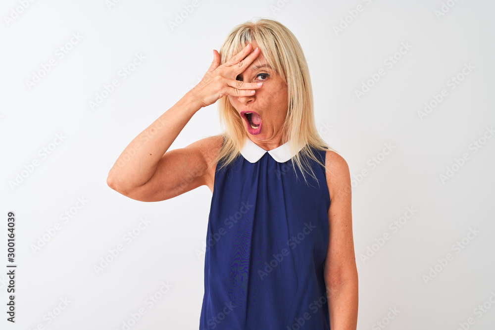 Middle age woman wearing elegant blue dress standing over isolated white background peeking in shock covering face and eyes with hand, looking through fingers with embarrassed expression. Middle age woman wearing elegant blue dress standing over isolated white background peeking in shock covering face and eyes with hand, looking through fingers with embarrassed expression.