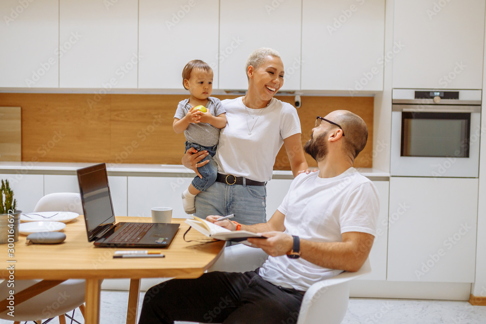 Fototapeta premium Happy family, Mother holding baby look at father. Friendly family in white clothing. Father sit with notebook
