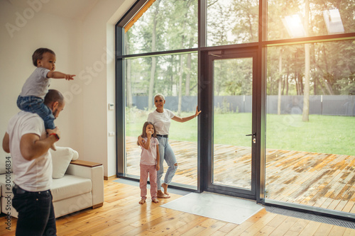 Parents with children look at panoramic window together, relaxing together, spending time at home. Modern house interior