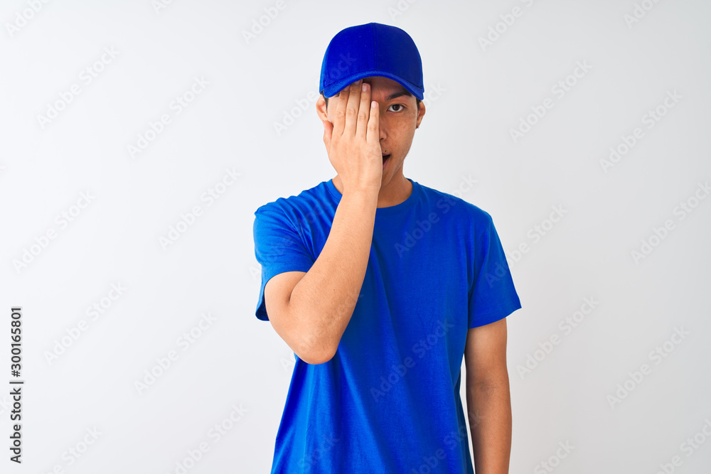 Chinese deliveryman wearing blue t-shirt and cap standing over isolated white background covering one eye with hand, confident smile on face and surprise emotion.