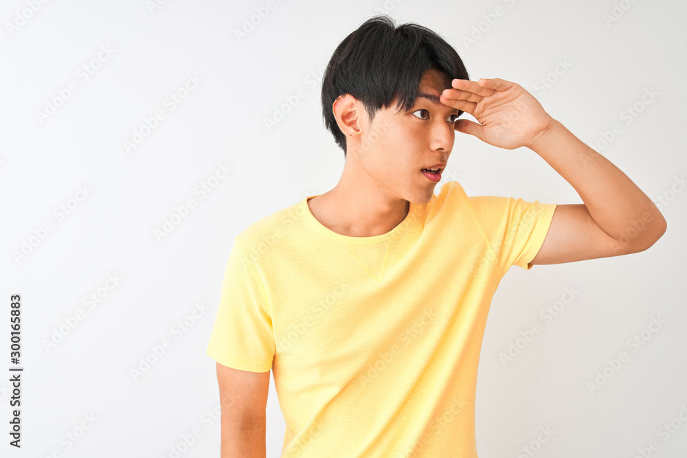 Chinese man wearing yellow casual t-shirt standing over isolated white background very happy and smiling looking far away with hand over head. Searching concept. Chinese man wearing yellow casual t-shirt standing over isolated white background very happy and smiling looking far away with hand over head. Searching concept.