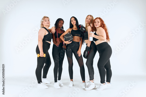 Five smiling multiethnic women wearing sportive leggins and topics holding fitness ball, boxing gloves and other things isolated over white background