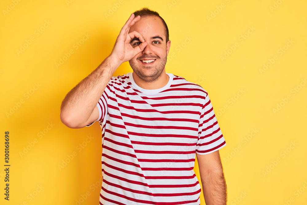 Young man wearing casual striped t-shirt standing over isolated yellow background doing ok gesture with hand smiling, eye looking through fingers with happy face. Young man wearing casual striped t-shirt standing over isolated yellow background doing ok gesture with hand smiling, eye looking through fingers with happy face.