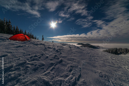 Red tourist tent  with a nightly lunar glow  on a snowy mountain peak  in the winter Ukrainian Carpathians  surrounded by foggy and frosty air 