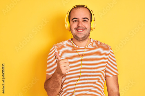 Young man listening to music using headphones standing over isolated yellow background doing happy thumbs up gesture with hand. Approving expression looking at the camera with showing success.