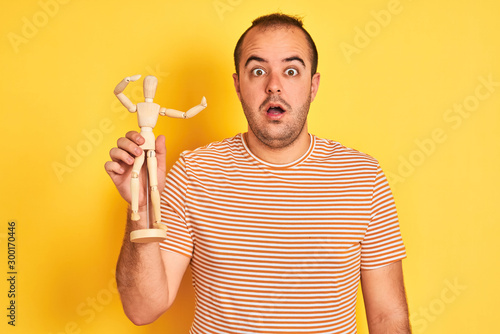 Young man holding figure of art dummy standing over isolated yellow background scared in shock with a surprise face, afraid and excited with fear expression