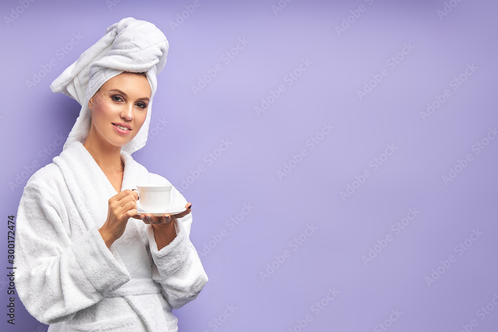 portrait of lovely woman with cup of tea or coffee, looking at camera. Female dressed in bathrobe and towel on head after shower. Isolated violet background portrait of lovely woman with cup of tea or coffee, looking at camera. Female dressed in bathrobe and towel on head after shower. Isolated violet background